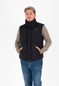 Zwarte gewatteerde vest met hoge kraag, rits aan de voorkant, twee zijzakken en klein logo op de linkerborst. Draagt over een beige langemouwen shirt.