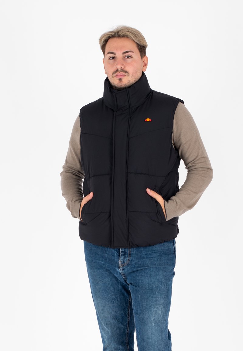 Zwarte gewatteerde vest met hoge kraag, rits aan de voorkant, twee zijzakken en klein logo op de linkerborst. Draagt over een beige langemouwen shirt.