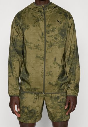 Homme portant une veste zippée à motif camouflage vert et olive foncé, avec un short assorti, debout devant un fond uni.