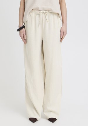 Femme portant un pantalon beige ample à cordon de serrage avec des jambes larges et des chaussures pointues foncées, debout devant un fond clair uni.