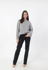 Lichtgrijze, geribbelde trui met brede mouwen, gecombineerd met zwarte, strakke broek. Model draagt beige open-teentjes schoenen en staat tegen een witte achtergrond.