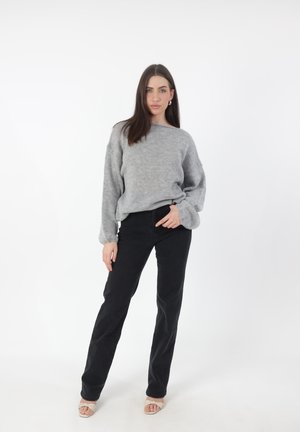 Lichtgrijze, geribbelde trui met brede mouwen, gecombineerd met zwarte, strakke broek. Model draagt beige open-teentjes schoenen en staat tegen een witte achtergrond.