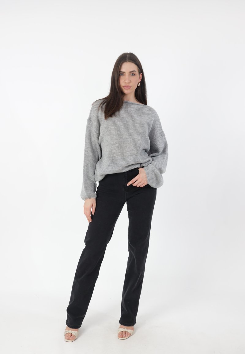 Lichtgrijze, geribbelde trui met brede mouwen, gecombineerd met zwarte, strakke broek. Model draagt beige open-teentjes schoenen en staat tegen een witte achtergrond.