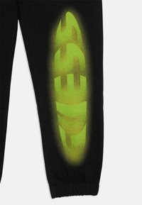 Pantalons de survêtement noirs avec un grand logo dégradé vert sur le côté. Le matériau est doux, avec des poignets élastiques aux chevilles.