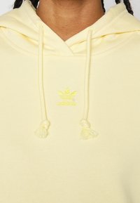 Žlutá mikina s kapucí má stahovací šňůrku s třásněmi, žebrovaný výstřih a vyšívané logo v odpovídající žluté barvě. Měkká bavlněná tkanina.