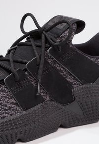 Black athletic shoe with a textured fabric upper, featuring thick laces, black suede accents, and a rubber outsole with a patterned design.