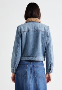 Jeansjacka med fårskinnskrage, kort design och klassisk blå tvättning. Baksidan visar sömmar och detaljerad sömnad.