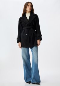 Sort bæltestøvle med en skarp krave, knapmanchetter og en skræddersyet pasform. Sammen med lyseblå udvidede jeans og sorte sko.