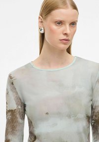 Naine sirgete blondide juustega, kes kannab hõbedaseid ümmargusi kõrvarõngaid ning läbi nähtava, pika varrukaga pluusi, millel on pehme abstraktne hall ja pruun muster.