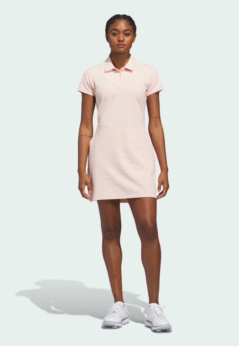 Kobieta stojąca w jasno różowej sukience polo i białych trampkach na jednolitym jasnym tle.
