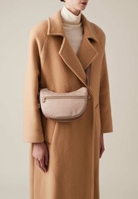 Cappotto di lana beige con revers, indossato sopra un dolcevita crema. Una borsa a tracolla in pelle rosa con una superficie testurizzata e una cerniera dorata è attaccata.