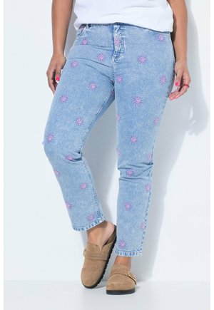 Jasnoniebieskie jeansy z różowym haftem słońca, noszone z beżowymi butami wsuwanymi i białą bluzką, ręce swobodnie opuszczone wzdłuż ciała.