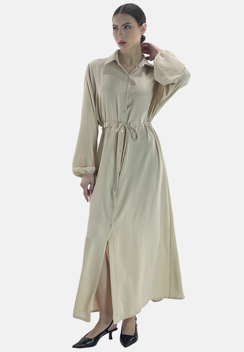 Robe chemise beige à manches longues avec un col, fermeture boutonnée sur le devant, taille cintrée, fente sur le côté et texture de tissu lisse. Portée avec des talons noirs.