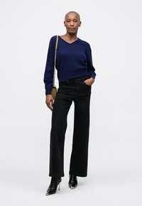 Pullover a V blu navy, jeans neri a gamba larga e stivaletti neri con tacco. Il modello porta una piccola borsa a tracolla verde oliva. Tessuto morbido.