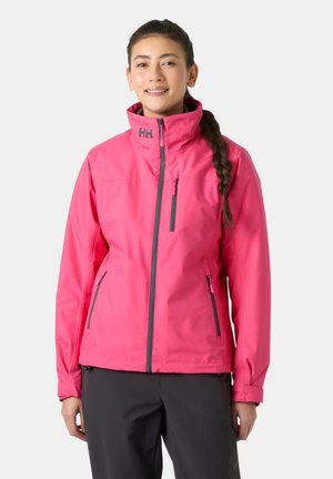 Mujer con cabello oscuro trenzado, sonriendo y vistiendo una chaqueta rosa brillante con cremallera y detalles en negro, y pantalones oscuros.