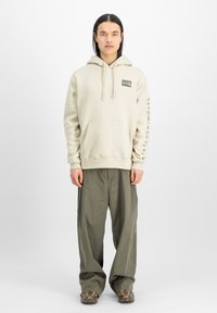 Beige huvtröja med framficka, med en rektangulär patch på bröstet och text på ärmen. Buren med löst sittande olivgröna byxor.