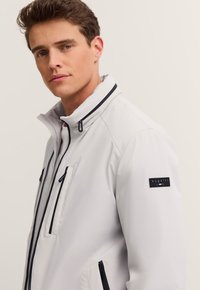 Hellgraue Jacke mit hohem Kragen, schwarzen Reißverschlussdetails, zwei Brusttaschen und einem "bugatti"-Label auf der linken Schulter. Glatte Textur.