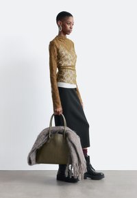 Frau in transparenter olivfarbener Bluse mit Blumenmuster, schwarzem Midikleid und schwarzen Stiefeln, die eine olivfarbene Handtasche mit einem grauen Schal vor einem schlichten weißen Hintergrund hält.