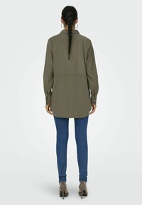 Camicia lunga verde oliva con colletto a punta, polsini abbottonati, orlo leggermente asimmetrico, abbinata a jeans skinny blu e tacchi nudi.