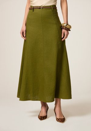Olijfgroene maxi-jurk met een licht uitlopende silhouette, voorzien van een bruine riem. Het model draagt leoprint hakken en armbanden.
