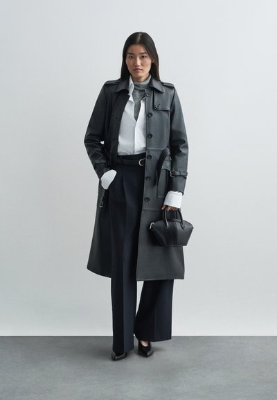 Trench coat in pelle nera con vita cintrata, grandi bottoni e vestibilità sartoriale. Abbinato a pantaloni larghi blu navy e una piccola borsetta nera.