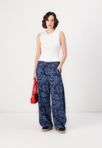 Top sans manches blanc, pantalon large à motifs floraux bleus avec une taille élastique, et un sac à main rouge. Modèle portant des chaussures noires avec des semelles beiges.