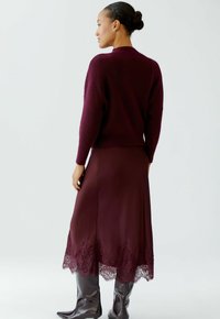 Burgunderfarbener Strickpullover mit lockerer Passform, kombiniert mit einem satinfarbenen burgunderfarbenen Rock mit Spitze und glänzenden knielangen braunen Stiefeln.