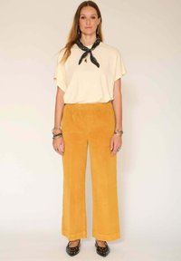 Haut couleur crème, ample, à manches courtes, associé à un pantalon large en velours côtelé jaune vif, et des chaussures noires à motifs.