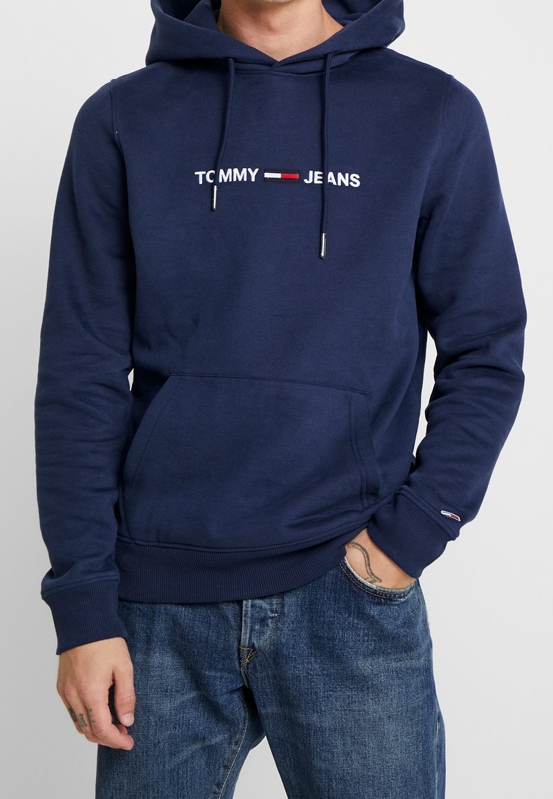 Man som bär en marinblå Tommy Jeans hoodie med en framficka och blå denimjeans, hand i fickan som visar en handledstatuering.