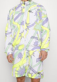 Wielokolorowy dres składający się z kurtki na zamek i krótkich spodenek, wykonany z lekkiego materiału z abstrakcyjnymi żółtymi, zielonymi i fioletowymi wzorami rozbłysków gwiazd.