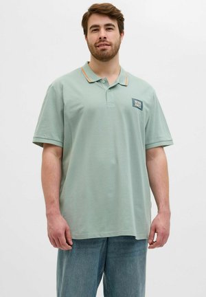 Svijetlozeleni polo majica s narančastim ovratnikom, s logotipom na lijevoj strani grudi. Kratki rukavi i ležeran kroj.