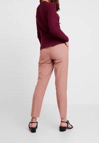 Frau trägt einen bordeauxfarbenen, strukturierten Pullover, pinke maßgeschneiderte Hose und schwarze Sandalen mit Absatz, steht mit einer Hand in der Tasche vor weißem Hintergrund.