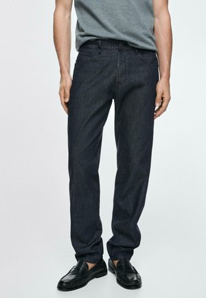 Mann trägt dunkelblaue gerade geschnittene Jeans, graues T-Shirt und schwarze Lederschlupfschuhe, steht mit entspannt an den Seiten hängenden Händen.