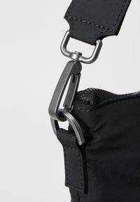 Sangle de sac en nylon noir avec un clip en métal. Présente une surface texturée et des coutures robustes, soulignant un élément de conception durable.