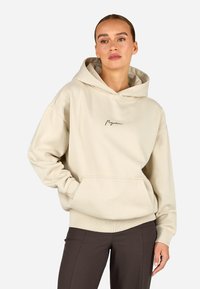 Μπεζ φούτερ με τσέπη καγκουρό και ραμμένες μανσέτες. Διαθέτει λογότυπο γραμμένο στο χέρι στο στήθος. Φτιαγμένο από μαλακό, υφασμένο ύφασμα.