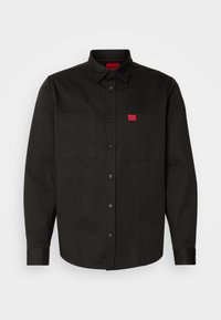 Chemise noire à manches longues avec boutons, col, deux poches poitrine et petite étiquette rouge "HUGO" sur la poche droite.