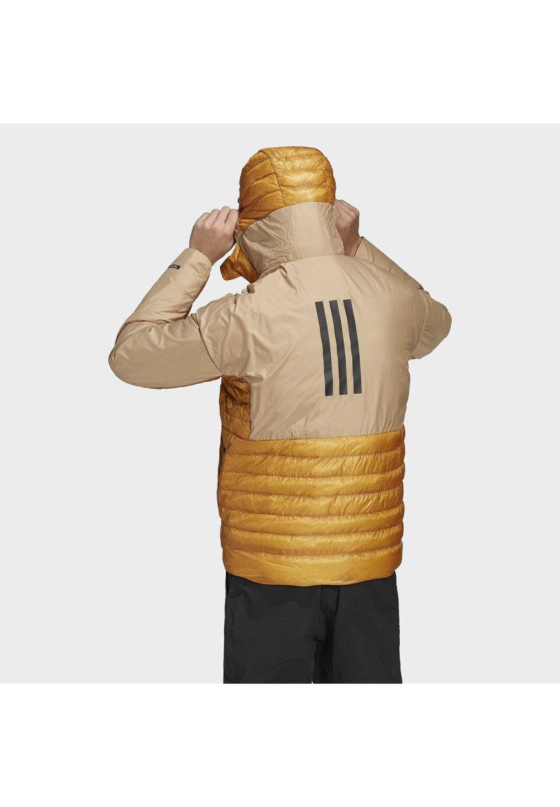 Person, der die Kapuze einer beigen und orangefarbenen Pufferjacke mit drei schwarzen Streifen auf dem Rücken anpasst, trägt schwarze Hosen.