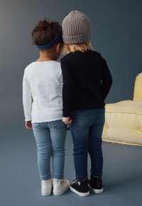 To børn set bagfra, den ene iført en hvid ribbet langærmet top og lyseblå jeans, den anden i en sort top og mørkeblå jeans.
