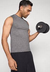 Γκρι αμάνικη αθλητική μπλούζα με εφαρμοστή σχεδίαση, φορεμένη από ένα άτομο που κρατάει έναν μαύρο αλτήρα με υφή και λευκό εσωτερικό.