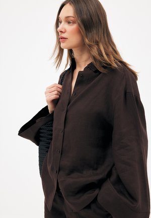 Giovane donna con capelli castano chiaro indossa una camicia marrone scuro a bottoni larga e porta una borsa nera intrecciata sulla spalla, guardando verso sinistra.