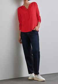 Rote Bluse mit langen Ärmeln, V-Ausschnitt und Knopfleiste, kombiniert mit dunkelblauen, weit geschnittenen Jeans und weißen Sneakers vor einem grauen Hintergrund.
