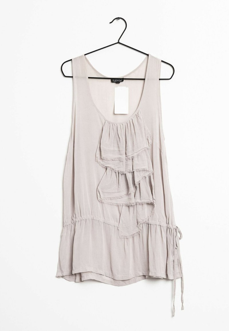 Blouse sans manches beige clair avec des volants superposés sur le devant, suspendue à un cintre noir sur fond blanc.