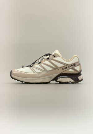 Beige en crèmekleurige Salomon trailrunning-schoen met zwarte trekkoordveters en een gestructureerde zool, zijaanzicht op een neutrale achtergrond.