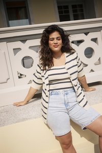 Mujer con cabello oscuro y ondulado se apoya en una barandilla de piedra, vestida con una camiseta a rayas y un cárdigan, junto con pantalones cortos de mezclilla claros, en un ambiente al aire libre soleado.