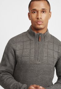 Grau gestrickter Pullover mit einem Viertel-Reißverschluss-Kragen, der ein strukturiertes Gittermuster und gerippte Bündchen aufweist. Das Material wirkt weich und warm.