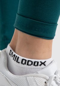 Turquoise leggings met een gladde textuur, voorzien van een gemarkeerde manchette. Witte sok met zwarte letters en een witte sneaker met gaasdetails.