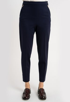 Granatowe spodnie o zwężanym kroju, o gładkiej fakturze, z zamkami błyskawicznymi w kolorze złota na przednich kieszeniach oraz dopasowanym paskiem w talii. Na nogach czarne mokasyny.