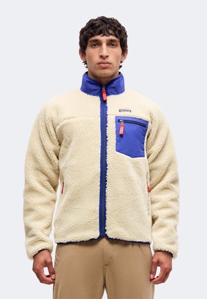 Jeune homme portant une veste polaire beige avec un col bleu et une poche poitrine, debout avec une expression neutre sur un fond uni.
