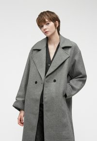 Pilkas vilnonis paltas su plačiu apykakle, dvigubos eigos priekiu ir dideliais juodais mygtukais. Laisvas siluetas, nuleistomis petnešomis ir šoninėmis kišenėmis.