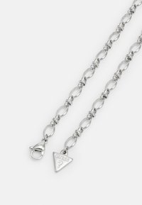 Zilveren ketting met ovale schakels, voorzien van een driehoekig label met "GUESS" in reliëf en een kreeftsluiting voor het sluiten.